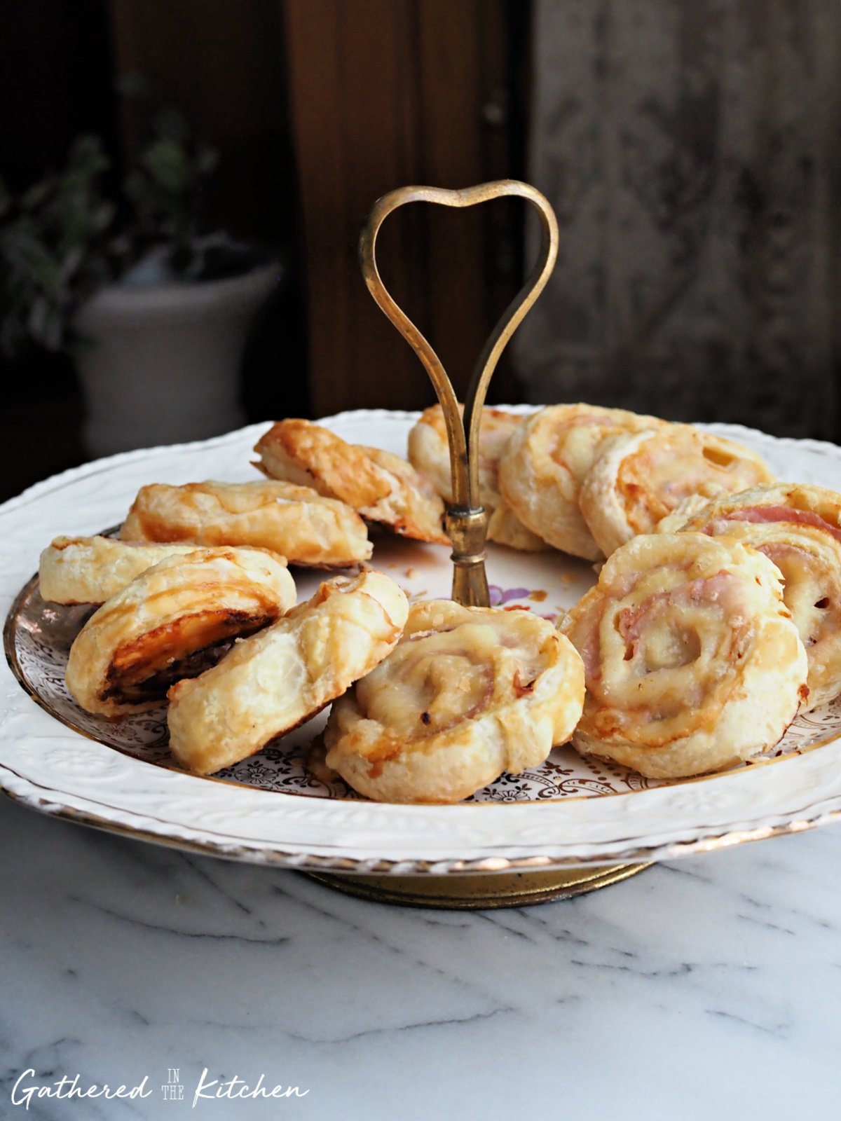 Flaky puff pastry pinwheel appetizers arranged on a white and gold tiered serving tray on a marble countertop. | Gathered In The Kitchen