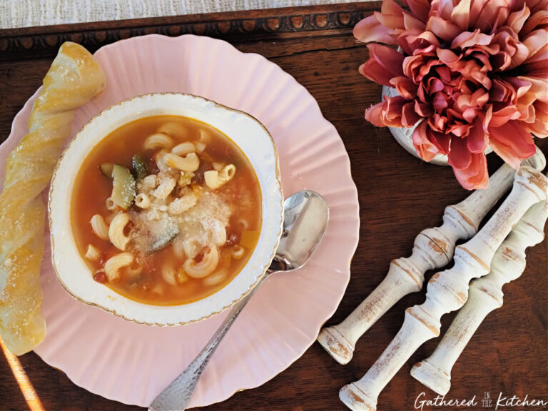 Bowl of vegetarian minestrone soup with pasta, zucchini, green beans, tomatoes, and beans served with bread on a pink plate