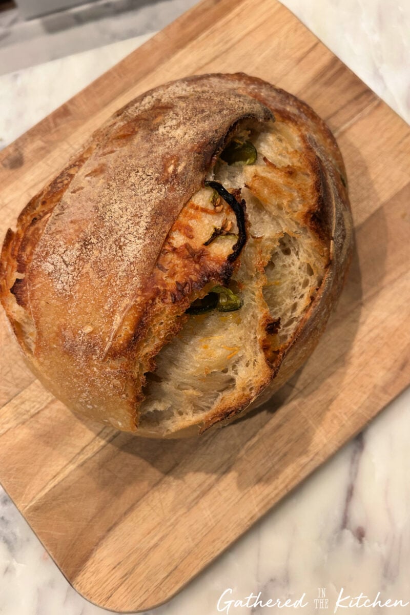 Baked jalapeño cheddar sourdough bread loaf with a golden crust and visible cheese and jalapeño pieces, partially sliced on a cutting board | Gathered In The Kitchen 