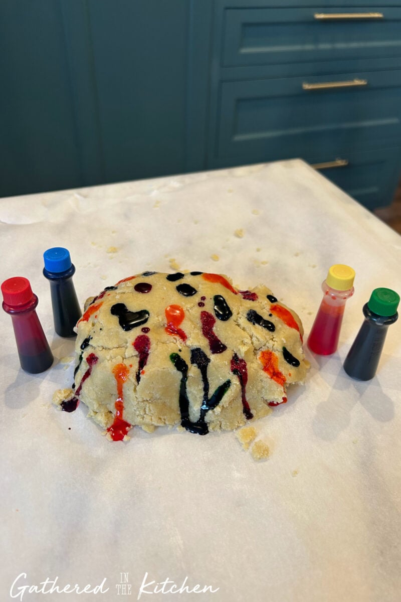 Sugar cookie dough with drops of red, blue, green, and yellow food coloring surrounded by small bottles on a marble countertop | Gathered In The Kitchen 