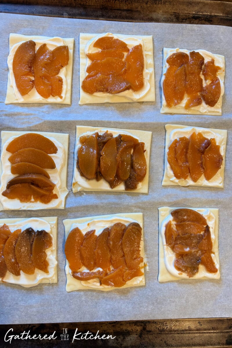 Sliced peaches layered on top of goat cheese filling on puff pastry squares before baking | Gathered In The Kitchen
