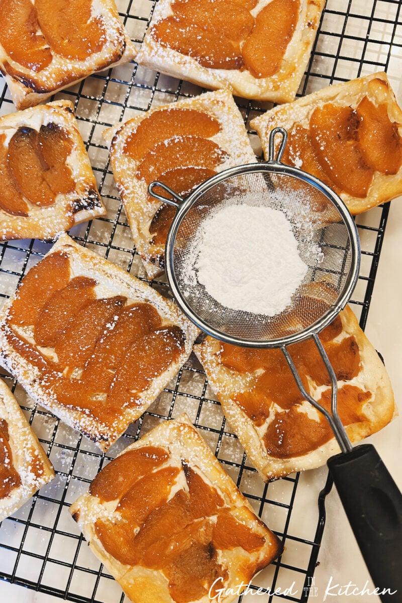 Powdered sugar being dusted over baked peach goat cheese danishes on a cooling rack | Gathered In The Kitchen