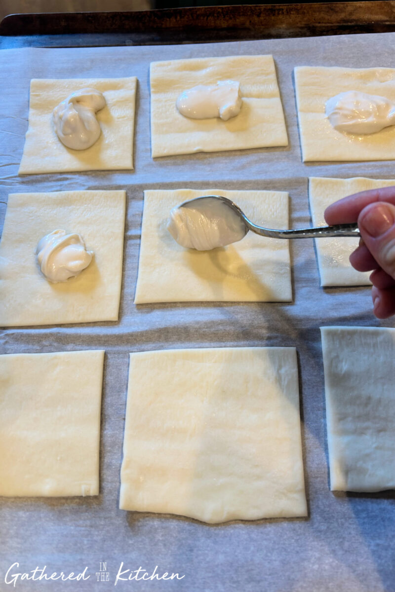 Spoon adding goat cheese filling onto puff pastry squares on a baking sheet | Gathered In The Kitchen 