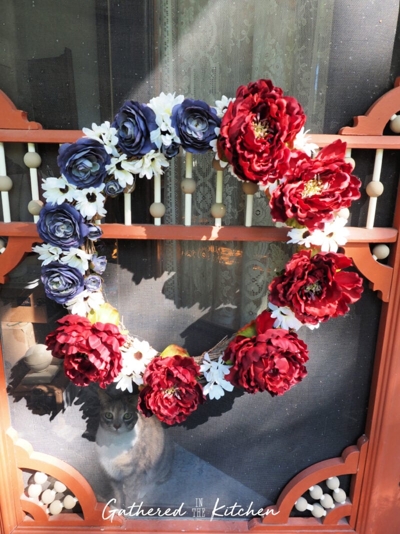 DIY patriotic floral wreath made from red, white, and blue thrift store faux flowers attached to a grapevine wreath form and displayed on a Victorian screen door for 4th of July decorating. | Gathered In The Kitchen 