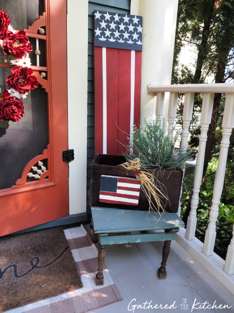 Patriotic porch decor featuring a DIY wooden American flag porch sitter sign, vintage wooden crate, faux greenery planter, and thrifted Americana decorations displayed beside a front door on a Victorian porch. | Gathered In The Kitchen 