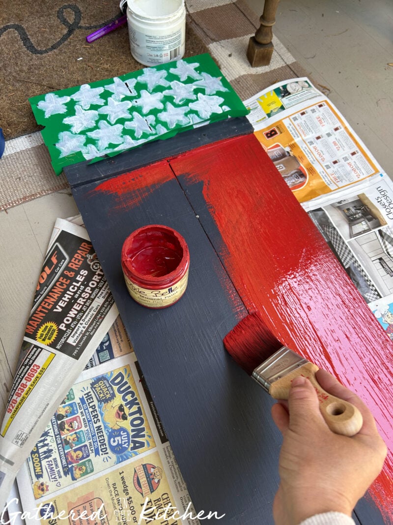Painting a thrifted wooden porch sitter sign red, white, and blue for DIY patriotic porch decor. This easy thrift flip transforms a secondhand wooden sign into a festive 4th of July porch decoration for a vintage-inspired front porch. | Gathered In The Kitchen 
