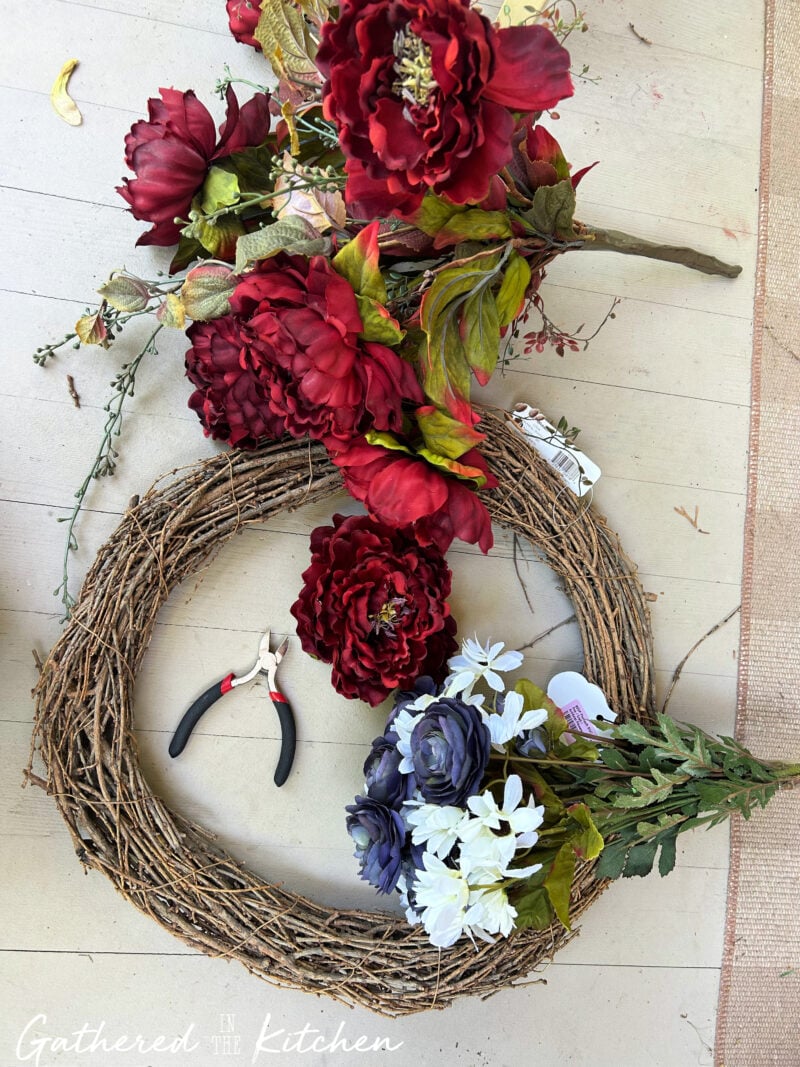 Supplies for a DIY patriotic wreath including a grapevine wreath form, red white and blue faux flowers, wire snips, and thrift store floral stems arranged on a porch floor before assembly. | Gathered In The Kitchen 