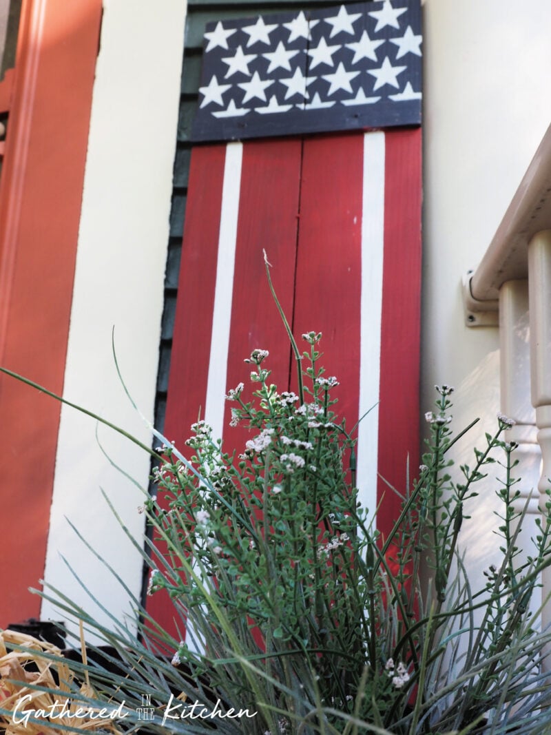 DIY wooden American flag porch sitter sign painted red, white, and blue and displayed with faux greenery as part of a thrifted patriotic porch decor arrangement for the 4th of July.