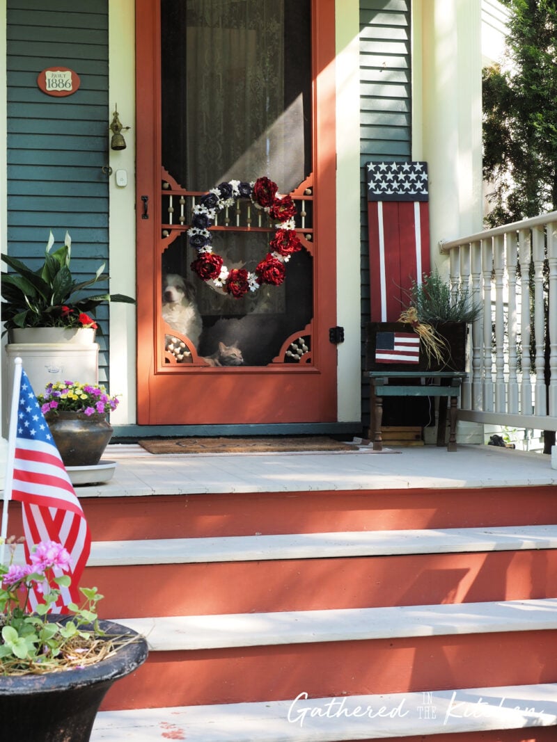 Finished patriotic front porch decorated with thrifted 4th of July decor including a DIY red, white, and blue floral wreath, wooden American flag porch sitter sign, vintage wooden crate, faux greenery, and American flags displayed on an 1886 Victorian home. | Gathered In The Kitchen 