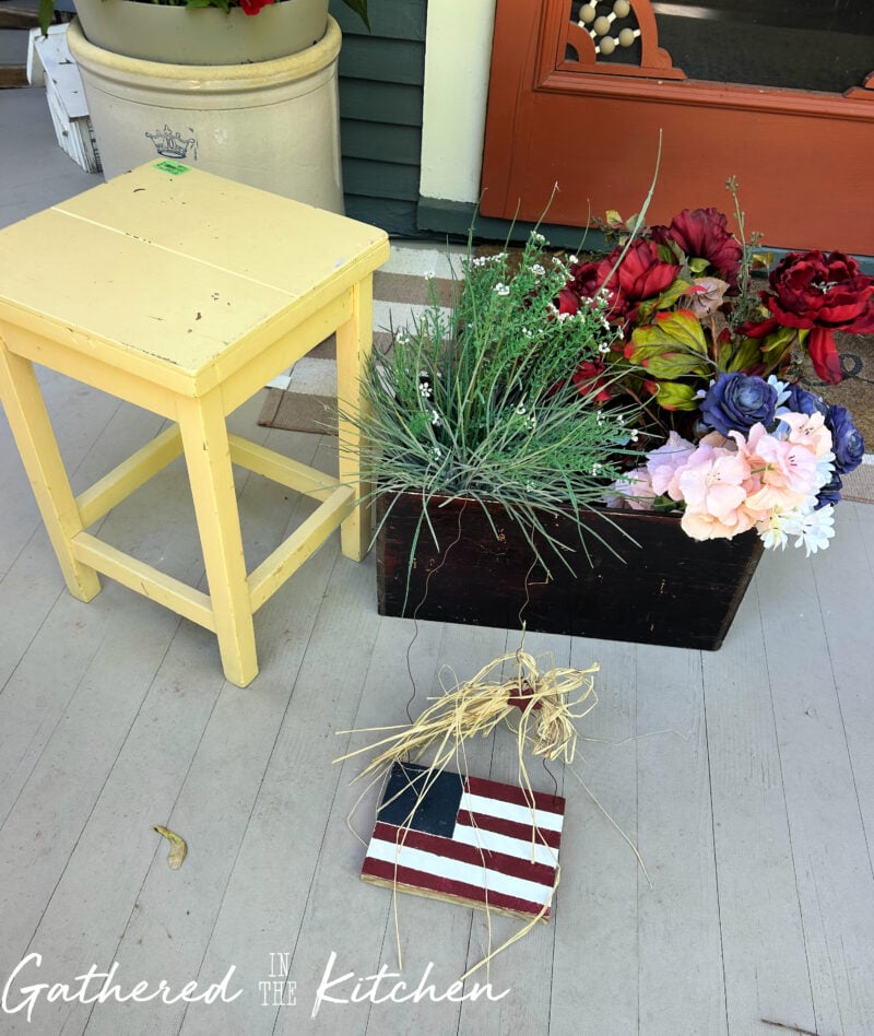 Collection of thrift store finds for DIY patriotic porch decor including a vintage wooden crate, faux flowers, greenery planter, Americana flag decor, and a yellow stool arranged on a Victorian front porch before styling. | Gathered In The Kitchen 