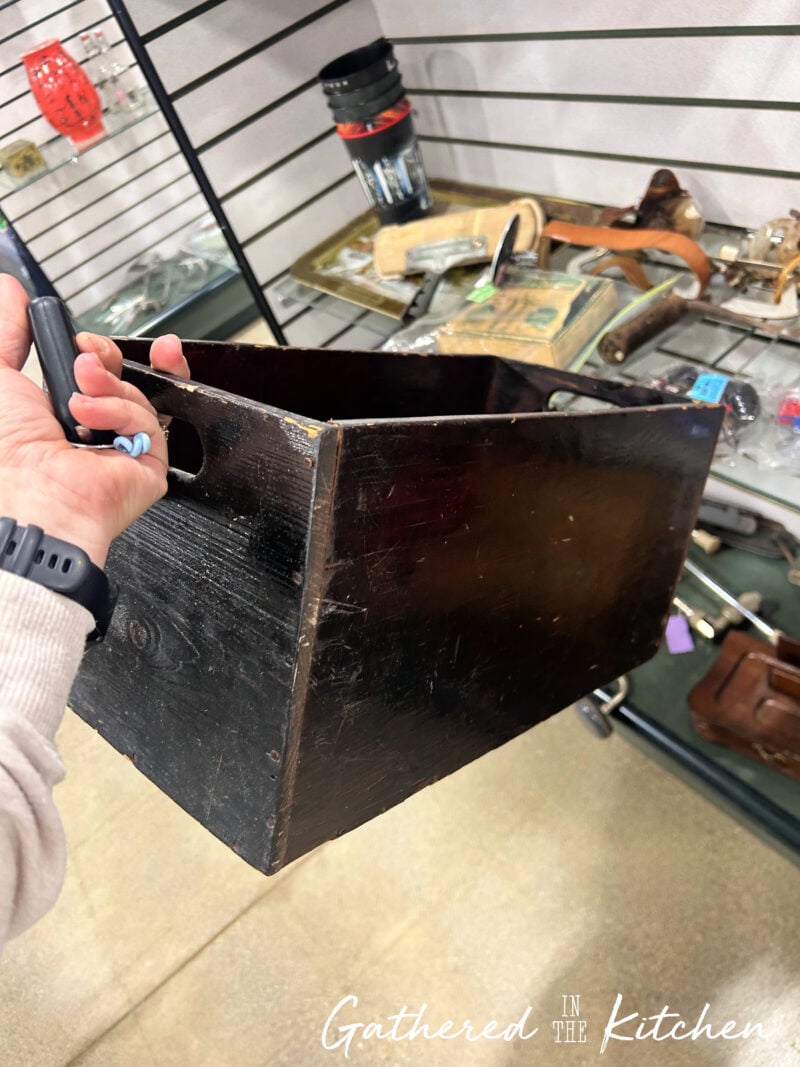 Holding a vintage wooden crate found at a thrift store that will be repurposed into patriotic porch decor for a 4th of July front porch display. The wooden box serves as a decorative base for thrifted Americana decorations and seasonal styling. | Gathered In The Kitchen 
