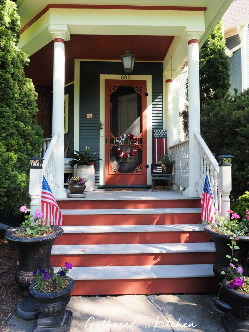 Front porch decorated for the 4th of July with thrifted patriotic decor, American flags, a DIY red white and blue wreath, vintage planters, and repurposed thrift store finds on an 1886 Victorian home. | Gathered In The Kitchen 