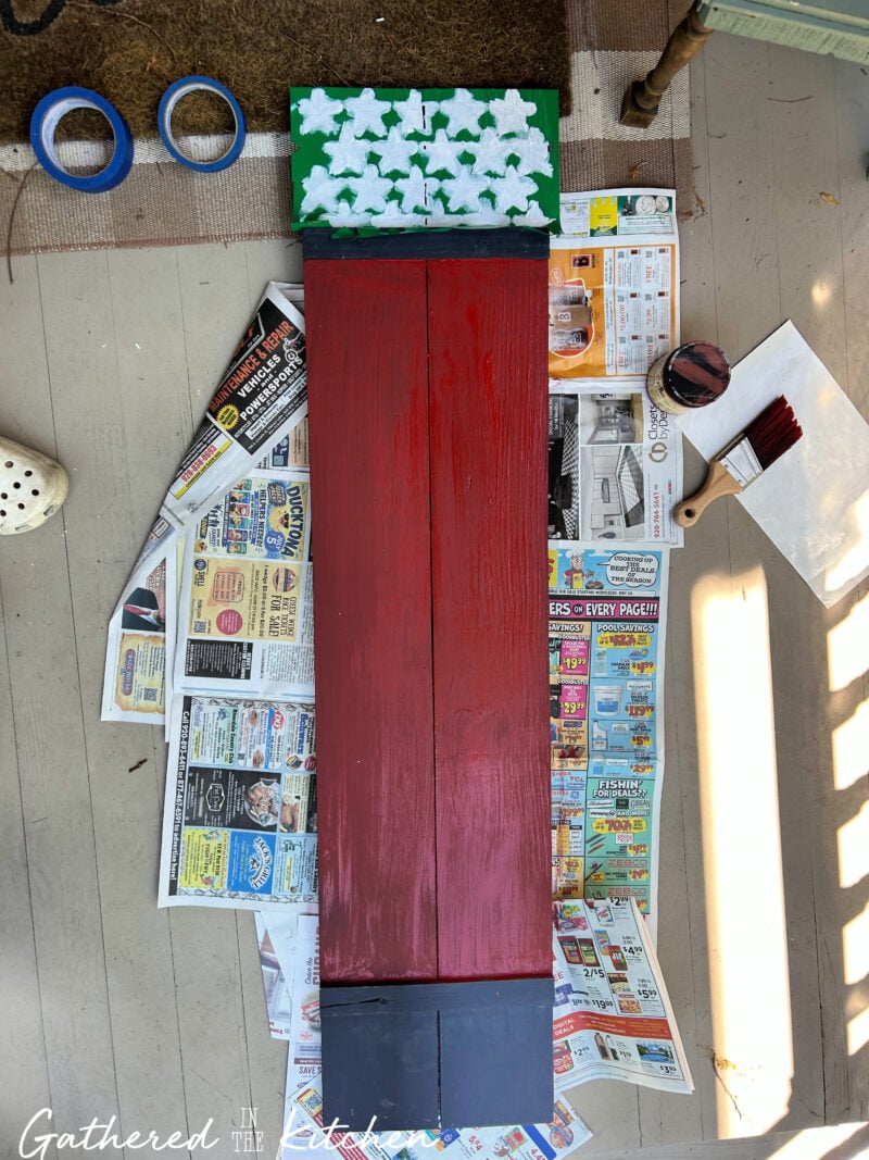 Patriotic wooden porch sign with painted red background and white star stencil section drying before adding white stripes to complete the Americana-inspired flag design | Gathered In The Kitchen