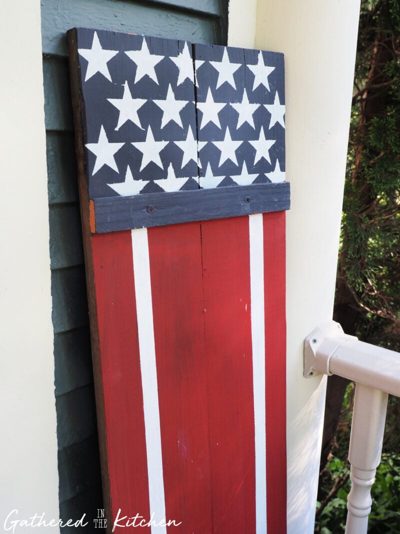 Finished DIY Americana wooden porch sign with white stars on a navy blue background and red and white flag-inspired stripes displayed on a front porch beside a railing | Gathered In The Kitchen
