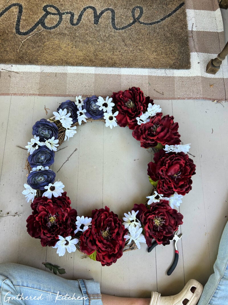 Red, white, and blue faux flowers arranged on a grapevine wreath form before being attached to create a DIY American flag-inspired 4th of July wreath. | Gathered In The Kitchen 