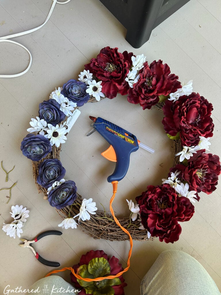 Red, white, and blue artificial flowers being attached to a grapevine wreath with a hot glue gun during a DIY patriotic wreath project. | Gathered In The Kitchen