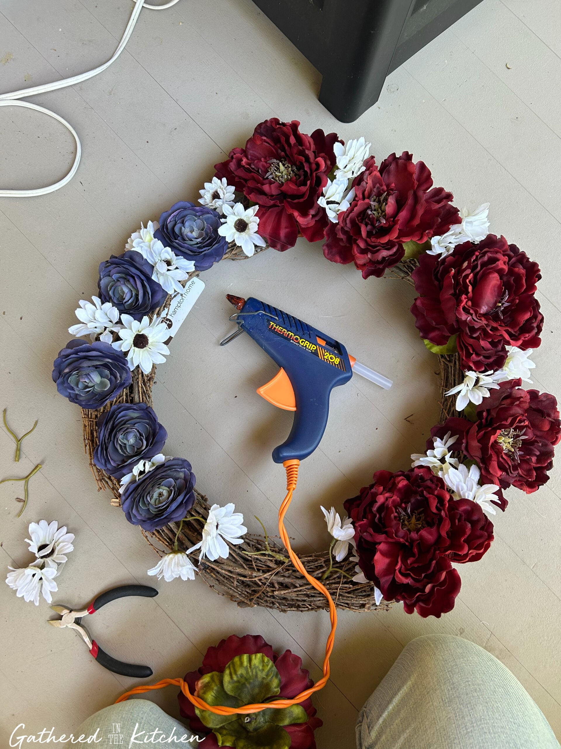 Red, white, and blue artificial flowers being attached to a grapevine wreath with a hot glue gun during a DIY patriotic wreath project. | Gathered In The Kitchen