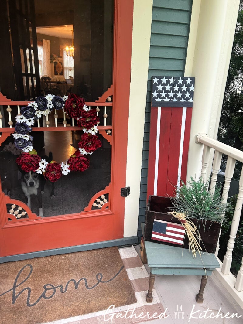 DIY Americana wooden porch sign displayed beside a front door with a patriotic floral wreath, vintage wooden crate, and repurposed porch stool on an 1886 Victorian home's front porch | Gathered In The Kitchen
