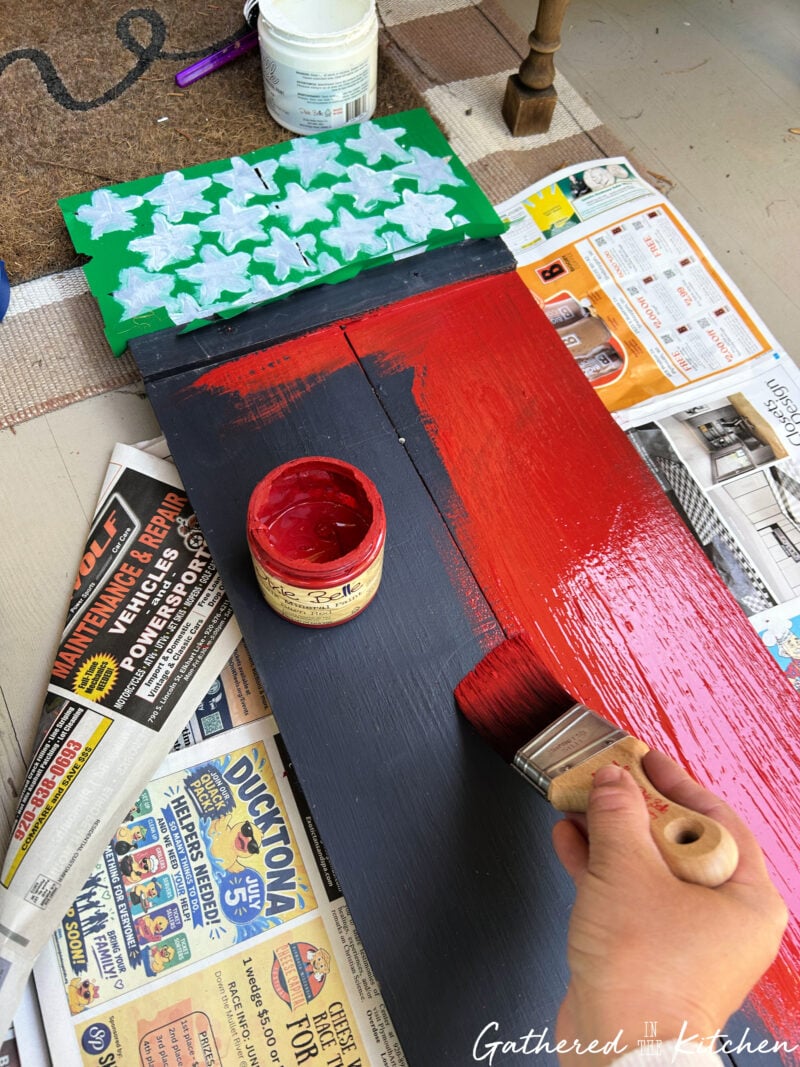Painting a bright red background onto a navy blue wooden porch sign to create the striped section of an Americana-inspired American flag design for patriotic front porch decor | Gathered In The Kitchen