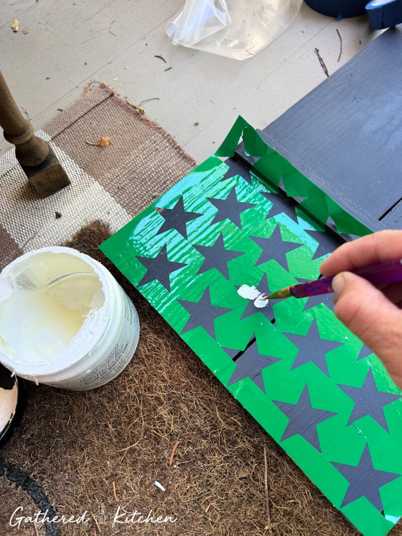 Painting white stars onto a navy blue wooden porch sign using custom vinyl stencils cut with a Silhouette Cameo to create a patriotic Americana-inspired front porch decoration | Gathered In The Kitchen
