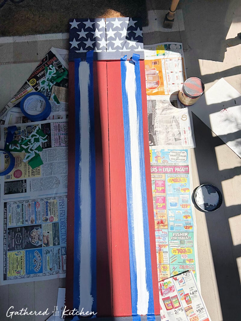 Painting white stripes between painter's tape lines on a patriotic wooden porch sign featuring white stars on a navy blue background and red flag-inspired stripes | Gathered In The Kitchen
