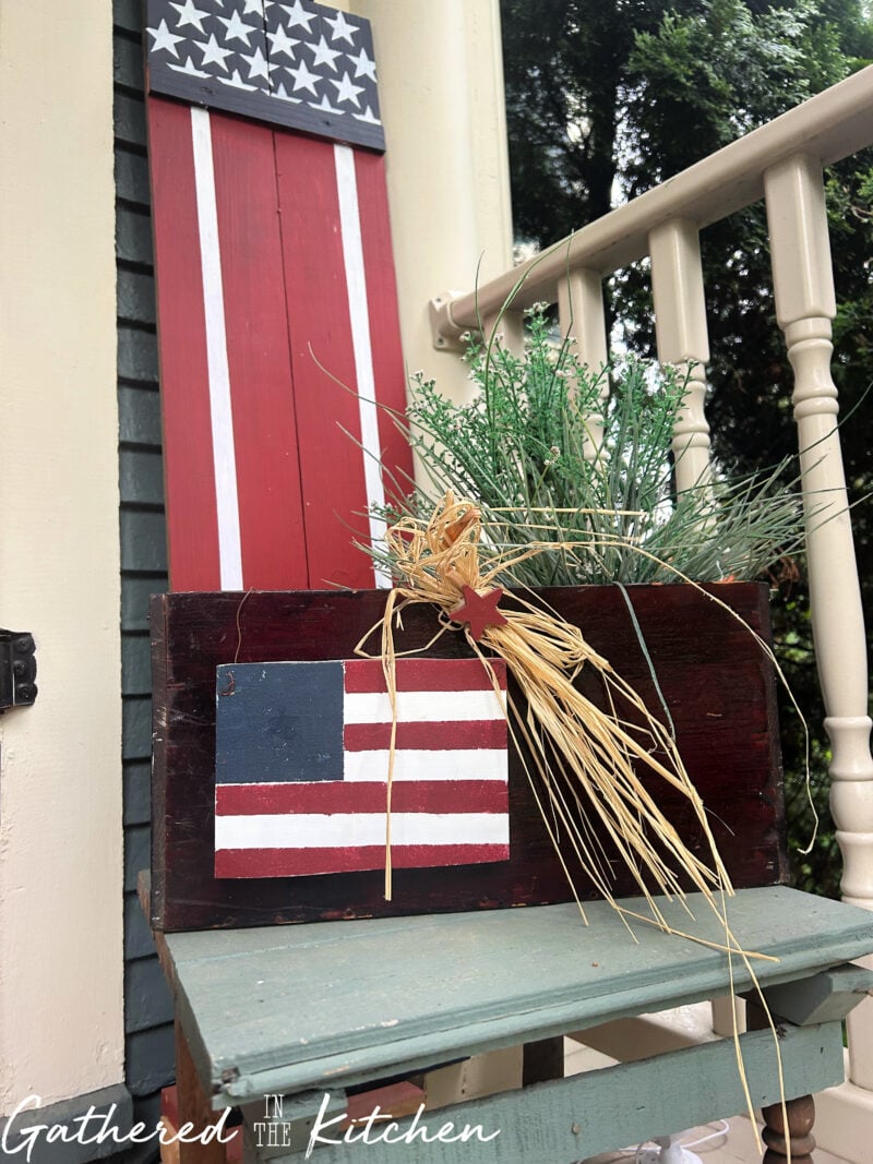 Patriotic porch display featuring a thrifted wooden box filled with faux greenery, a wooden American flag accent, and a handmade Americana porch sign for 4th of July decorating. | Gathered In The Kitchen 