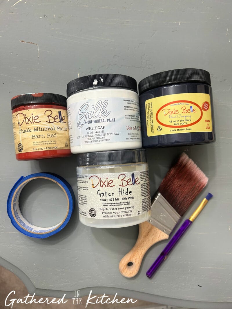 Paint supplies including red, white, and navy blue paint, painter's tape, paint brushes, and topcoat used to create a DIY Americana wooden porch sign with a patriotic flag-inspired design | Gathered In The Kitchen
