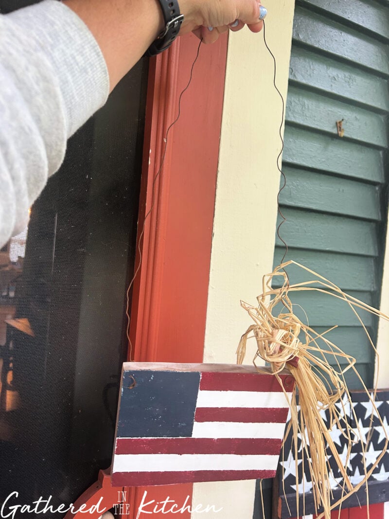 Hand holding a thrifted wooden American flag decoration with a wire hanger and raffia bow before adding it to a patriotic porch display for the 4th of July. | Gathered In The Kitchen 