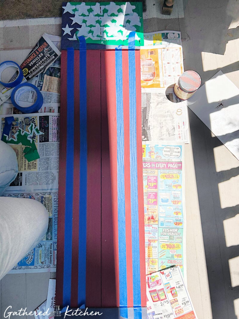 Blue painter's tape applied to a wooden porch sign to create evenly spaced white flag stripes over a red background for a DIY Americana-inspired patriotic porch decoration | Gathered In The Kitchen
