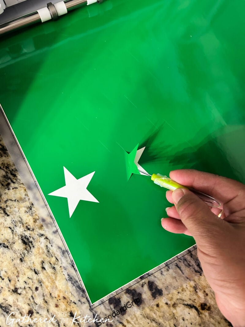 Hand weeding a star-shaped vinyl stencil cut with a Silhouette Cameo on green vinyl for a DIY patriotic wooden porch sign featuring an Americana-inspired American flag design. | Gathered In The Kitchen